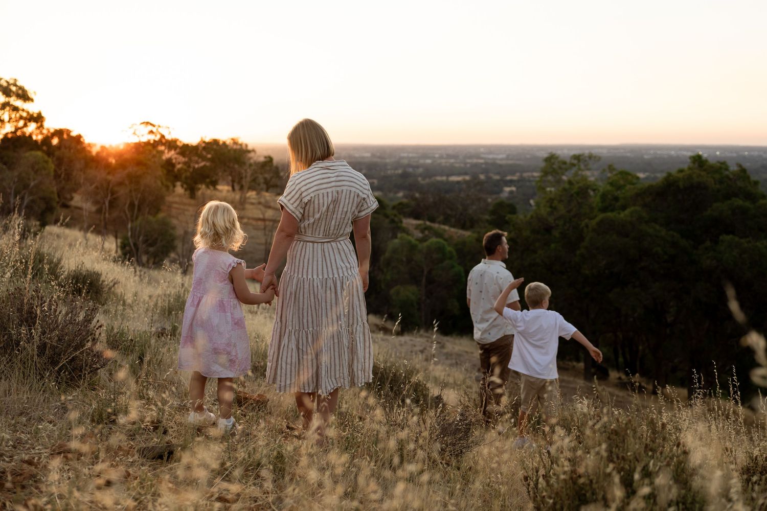 Family of four holding hands at golden sunset on grassy hill overlooking scenic landscape.