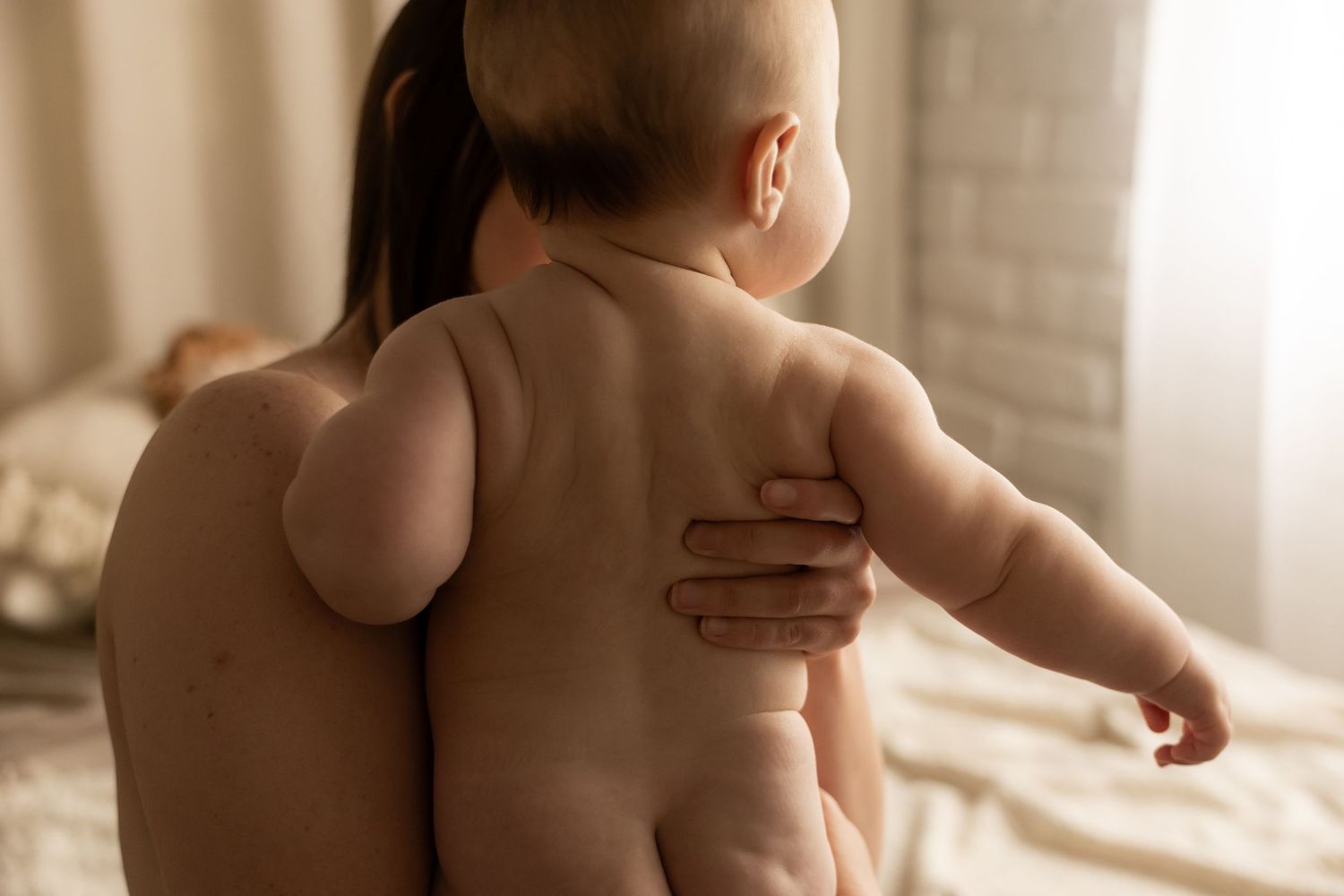 A mother holds her naked baby against her bare back, sharing a tender bonding moment in a softly lit bedroom.