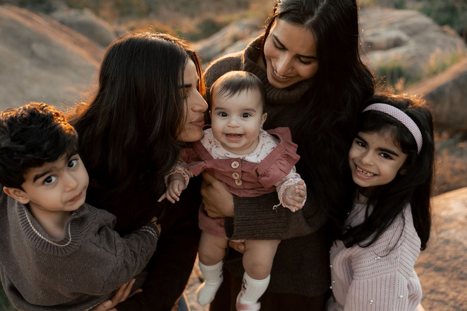 Family portrait of two women with three children, including a smiling baby in a pink dress, outdoors at golden hour in the Perth hills