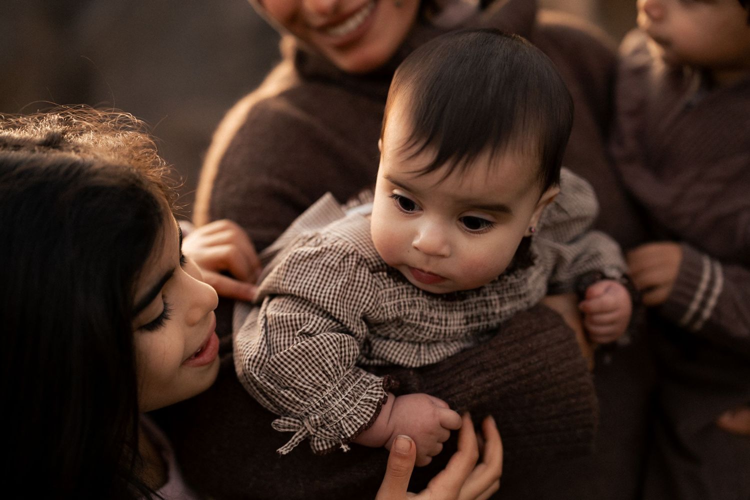 A baby in a checkered outfit being held by her mother women in warm brown tones.