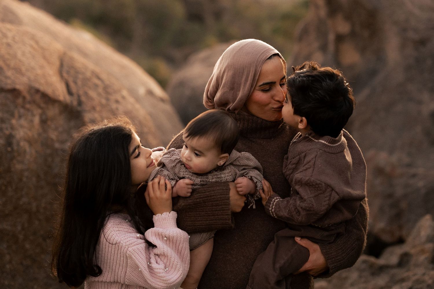 A mother in a hijab tenderly embraces her three young children outdoors among rocky terrain in warm natural light.