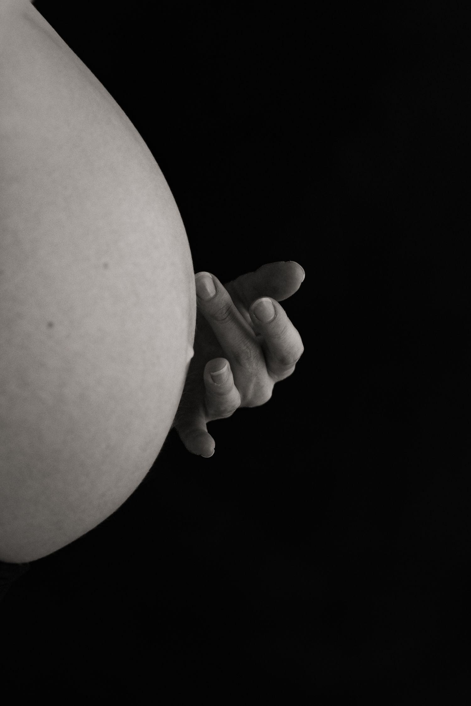 Black and white photo of a pregnant belly with a small baby hand reaching around the side against dark background.