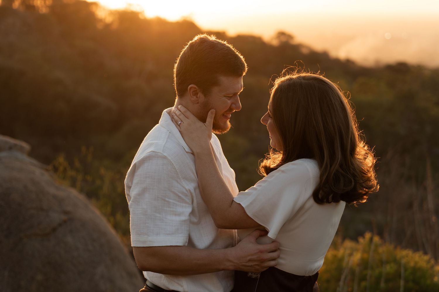 Couple embracing at golden hour sunset on a hilltop, sharing a romantic moment with warm glowing light behind them.
