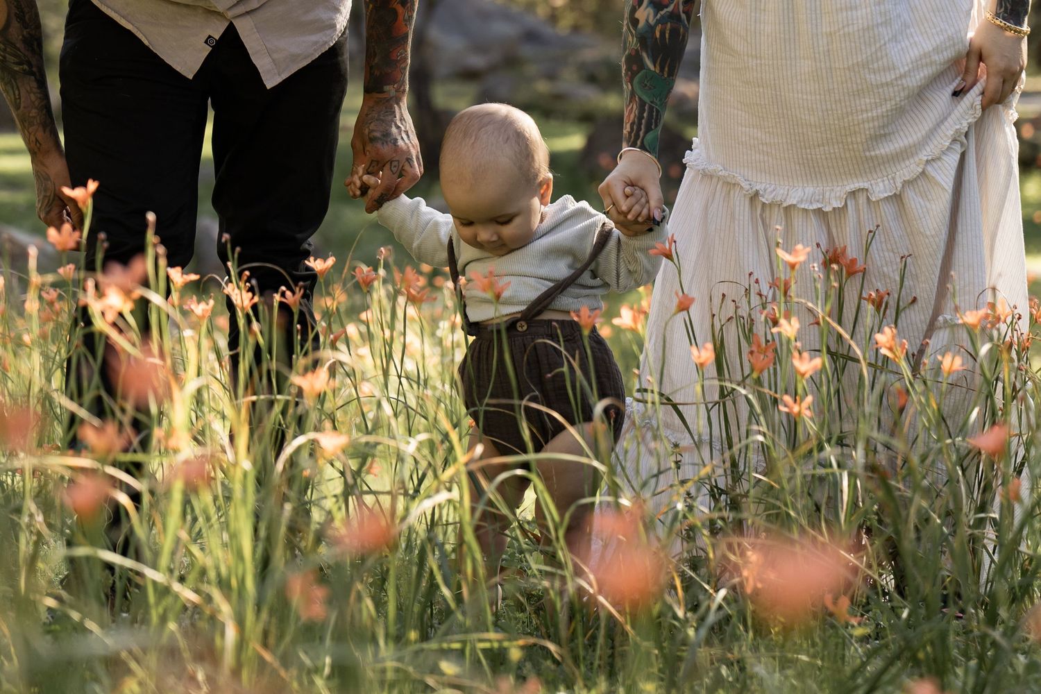 Baby taking first steps through wildflowers, supported by parents hands in a sunny garden.