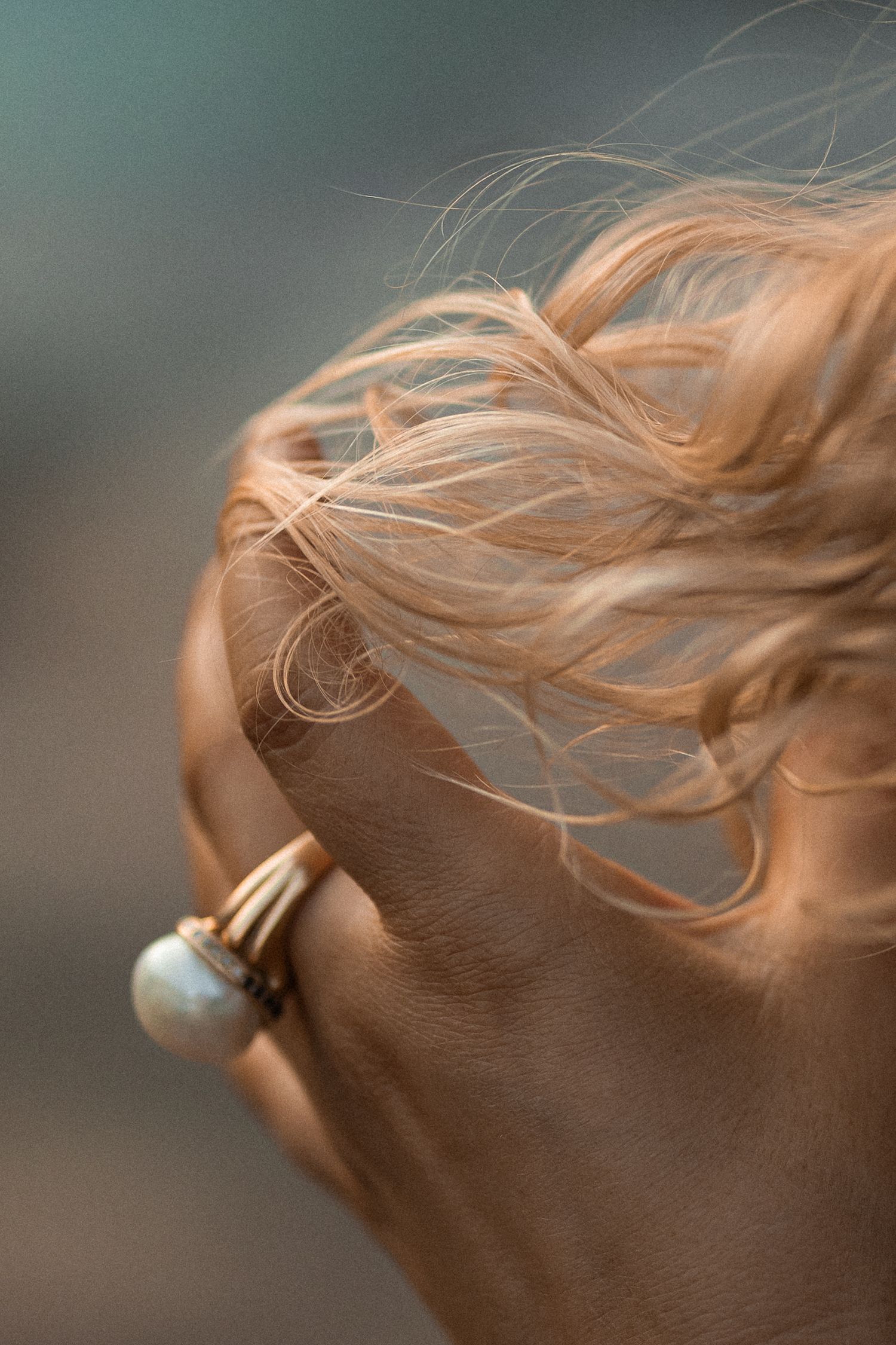 Close-up of a hand with a pearl ring holding windswept blonde hair against a blurred teal background.