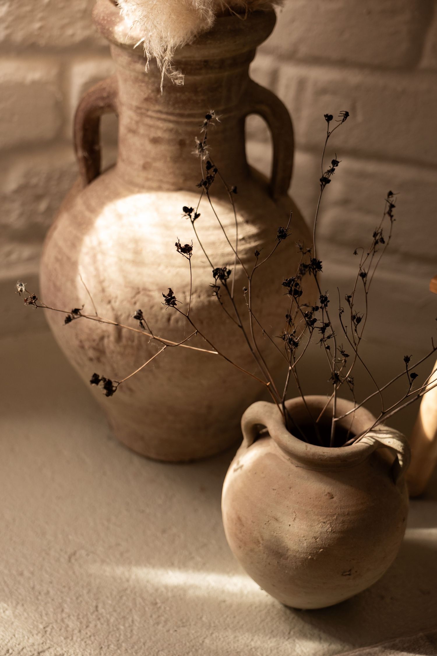 Rustic terracotta clay vases with dried wildflowers in warm sunlight against a white brick wall.