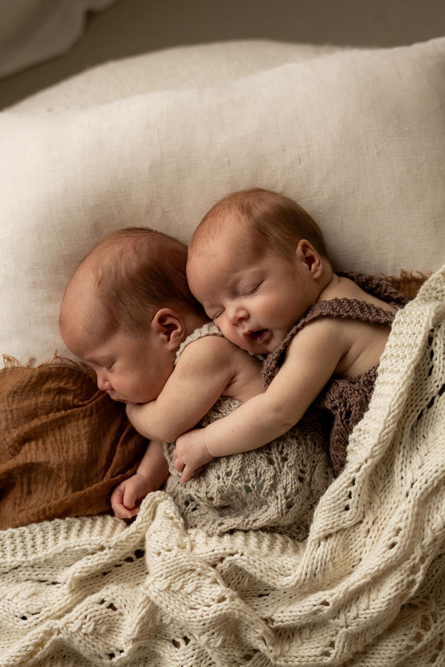 Two sleeping newborn twins cuddled together on soft cream and brown knit blankets in a cozy basket.