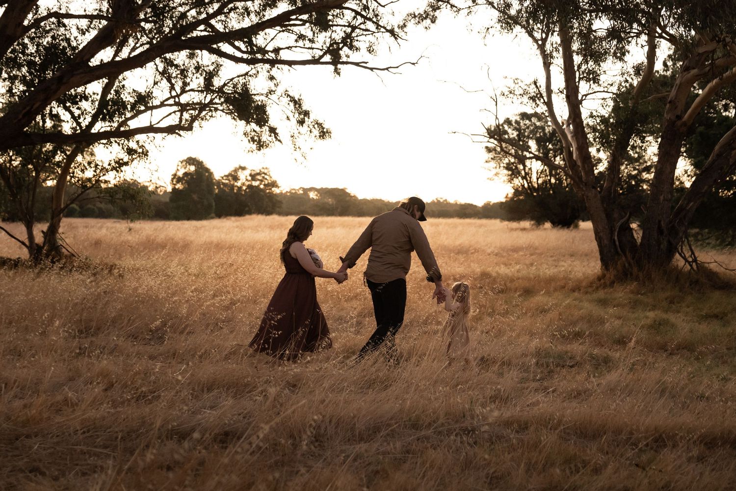 Family of three holding hands walking through golden grass field at sunset near trees.