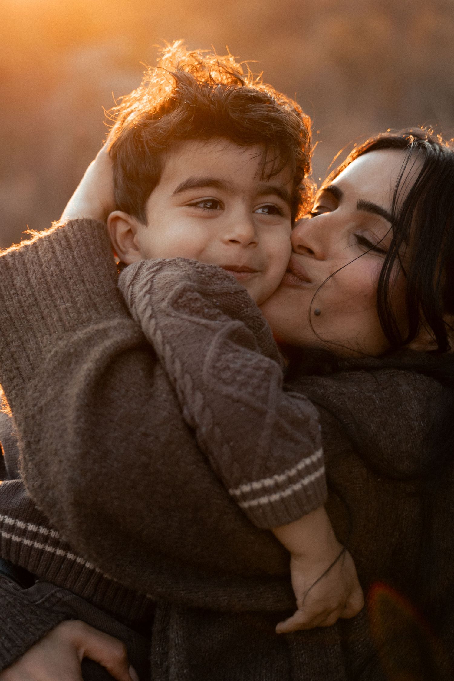 A mother tenderly kisses her young son's cheek during a warm golden sunset, both wearing cozy brown sweaters.