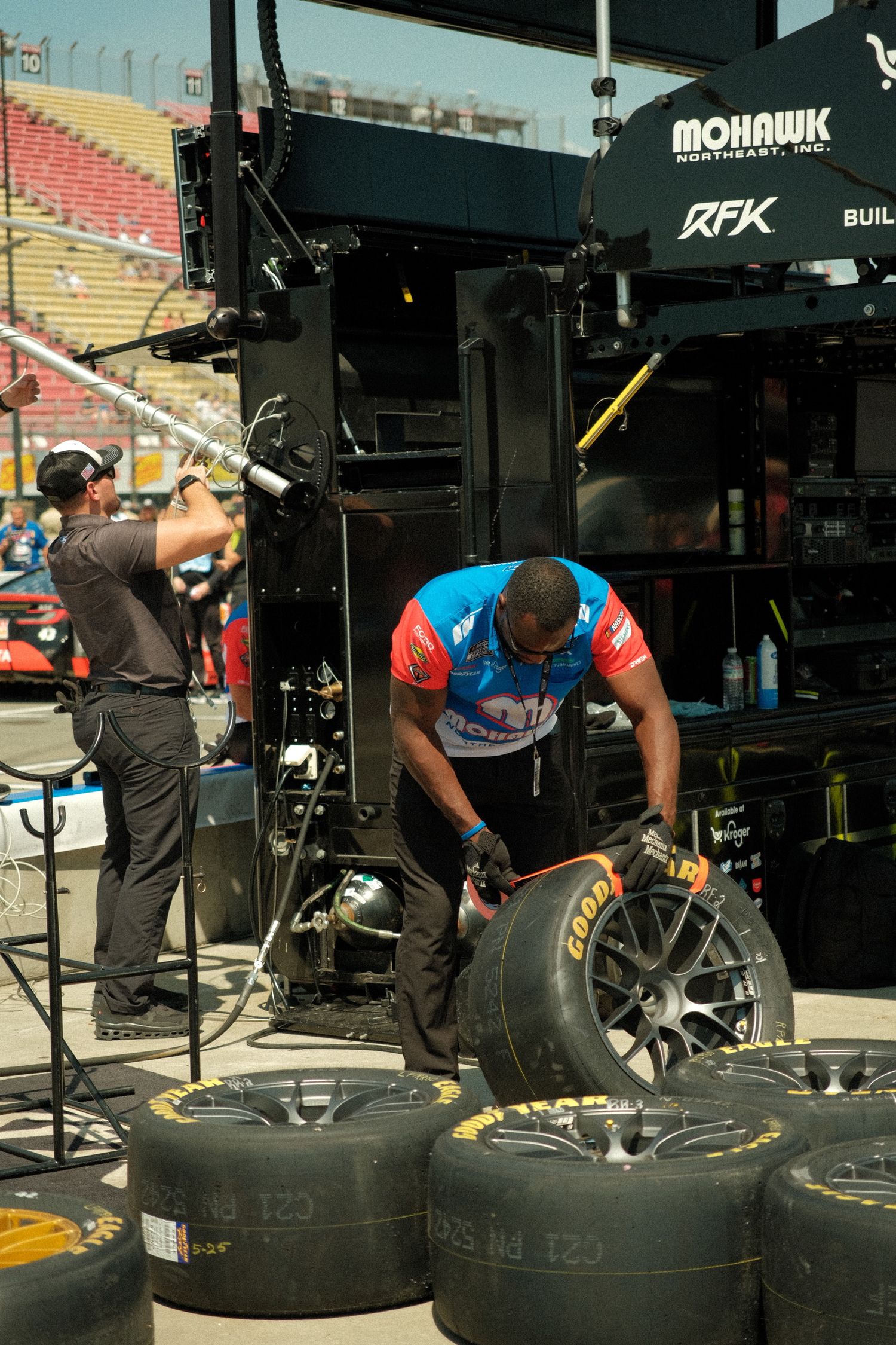 RFK Racing crew member handles Goodyear tires in pit garage area during NASCAR race day preparations.