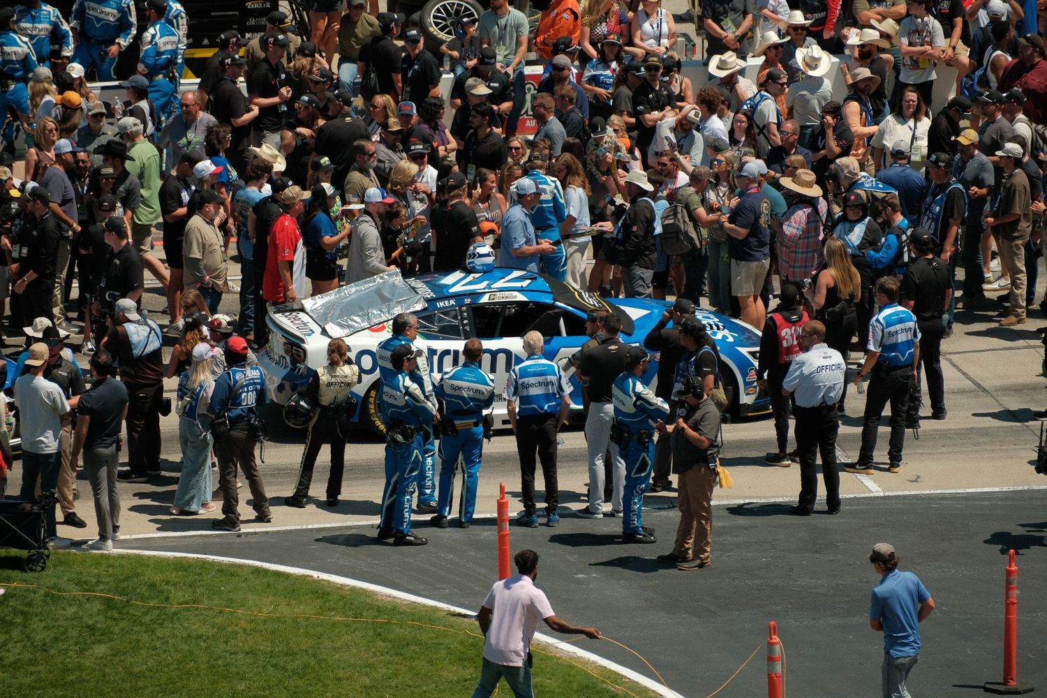 NASCAR pit crew members surround a blue race car amid a large crowd of spectators at a racing event.