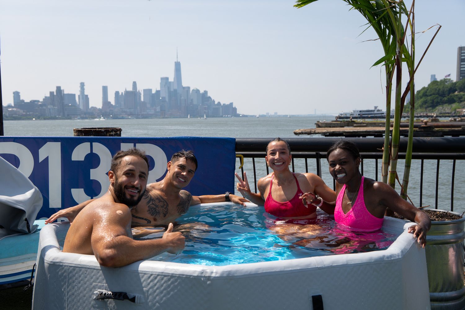 Four friends smiling in a hot tub with the New York City skyline visible in the background.