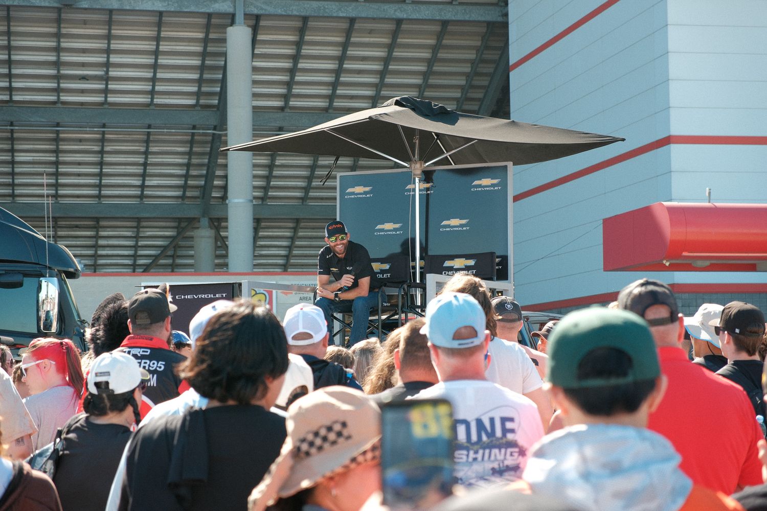NASCAR driver addresses a large crowd of fans gathered outside a racing facility at an outdoor fan event.