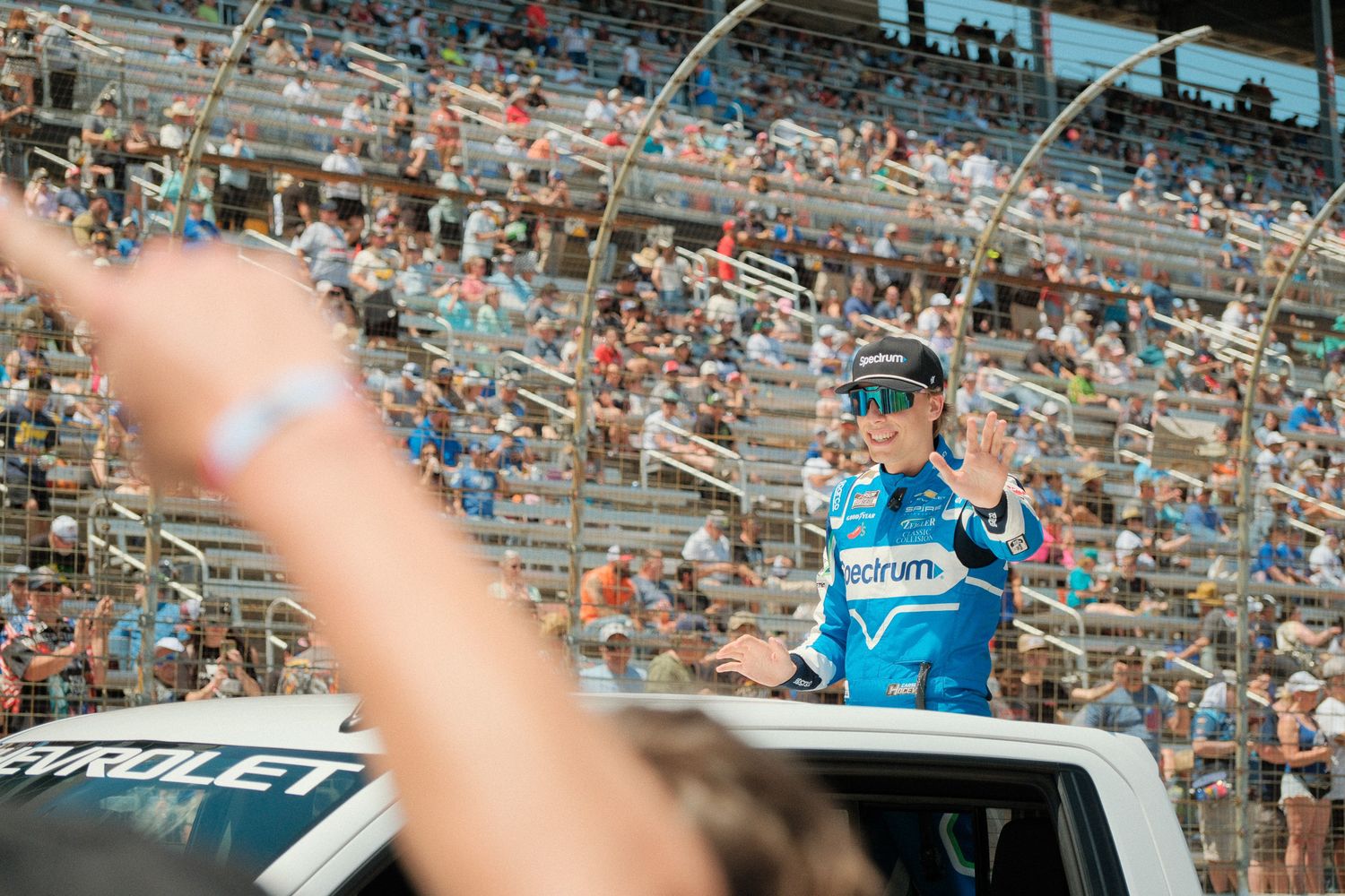 NASCAR driver waves to cheering crowd from vehicle during pre-race parade at packed speedway.