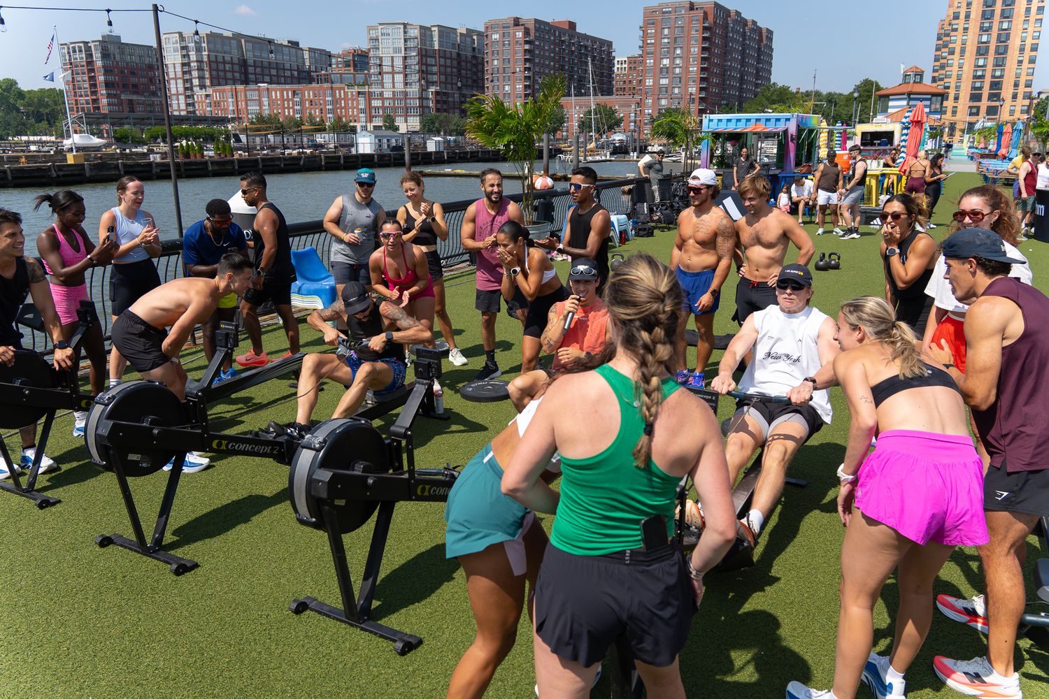 Large outdoor group fitness class with rowing machines and participants on waterfront turf.