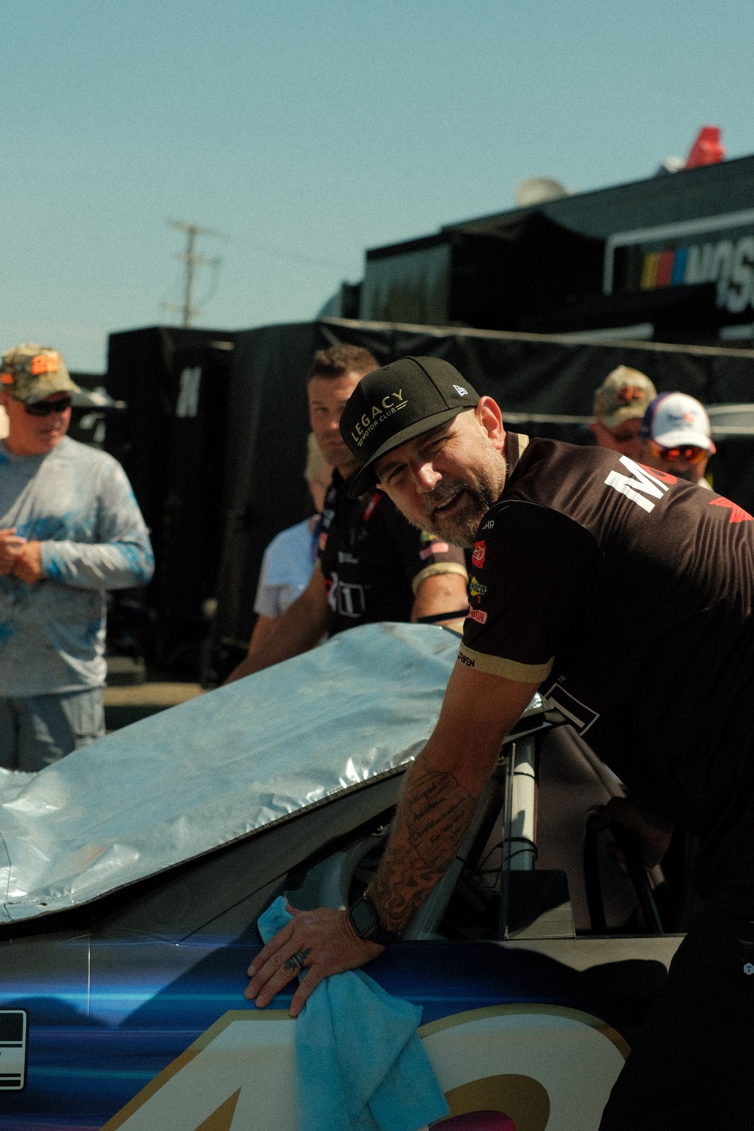 A tattooed NASCAR crew member in a Legacy Motor Club cap leans over a number 42 John Hunter Nemechek race car in the garage at Watkins Glen.