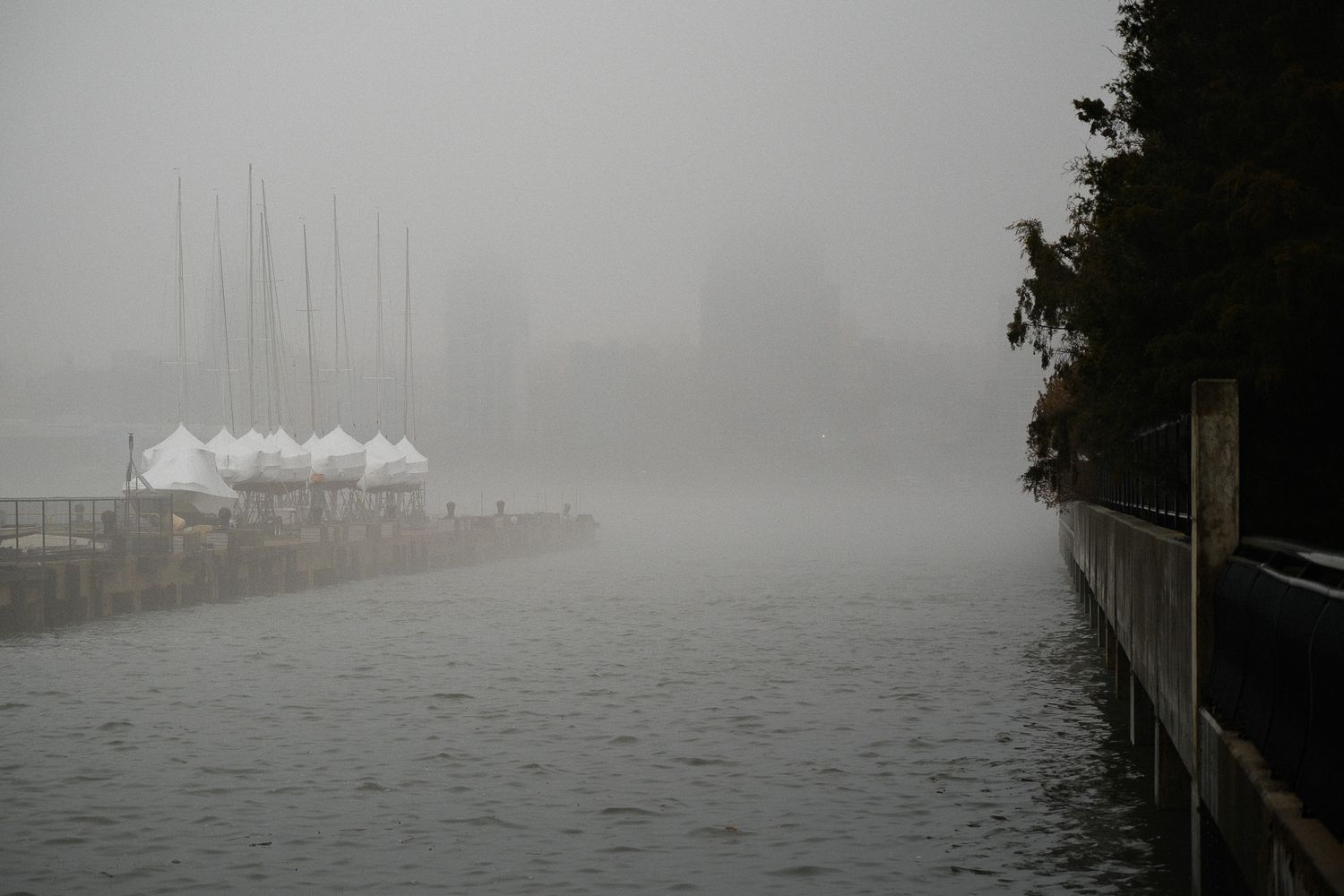 Misty foggy marina with covered sailboats along waterfront path disappearing into thick morning haze in Hoboken, NJ.