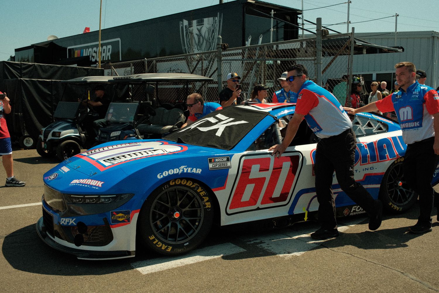 NASCAR #60 blue and white sponsored race car being pushed by crew members in pit lane.