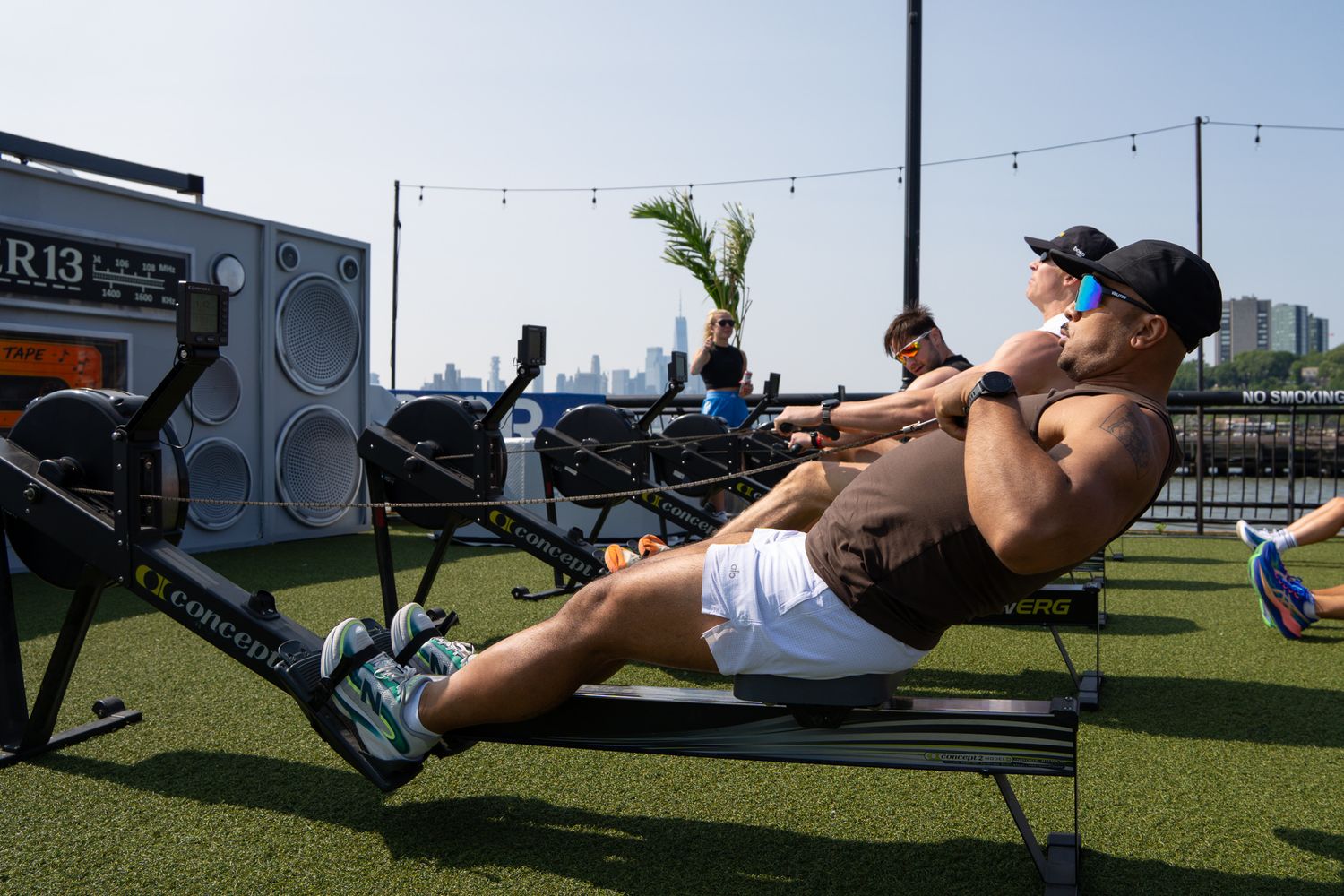 Athletes rowing on Concept2 machines outdoors on a rooftop with city skyline in background.