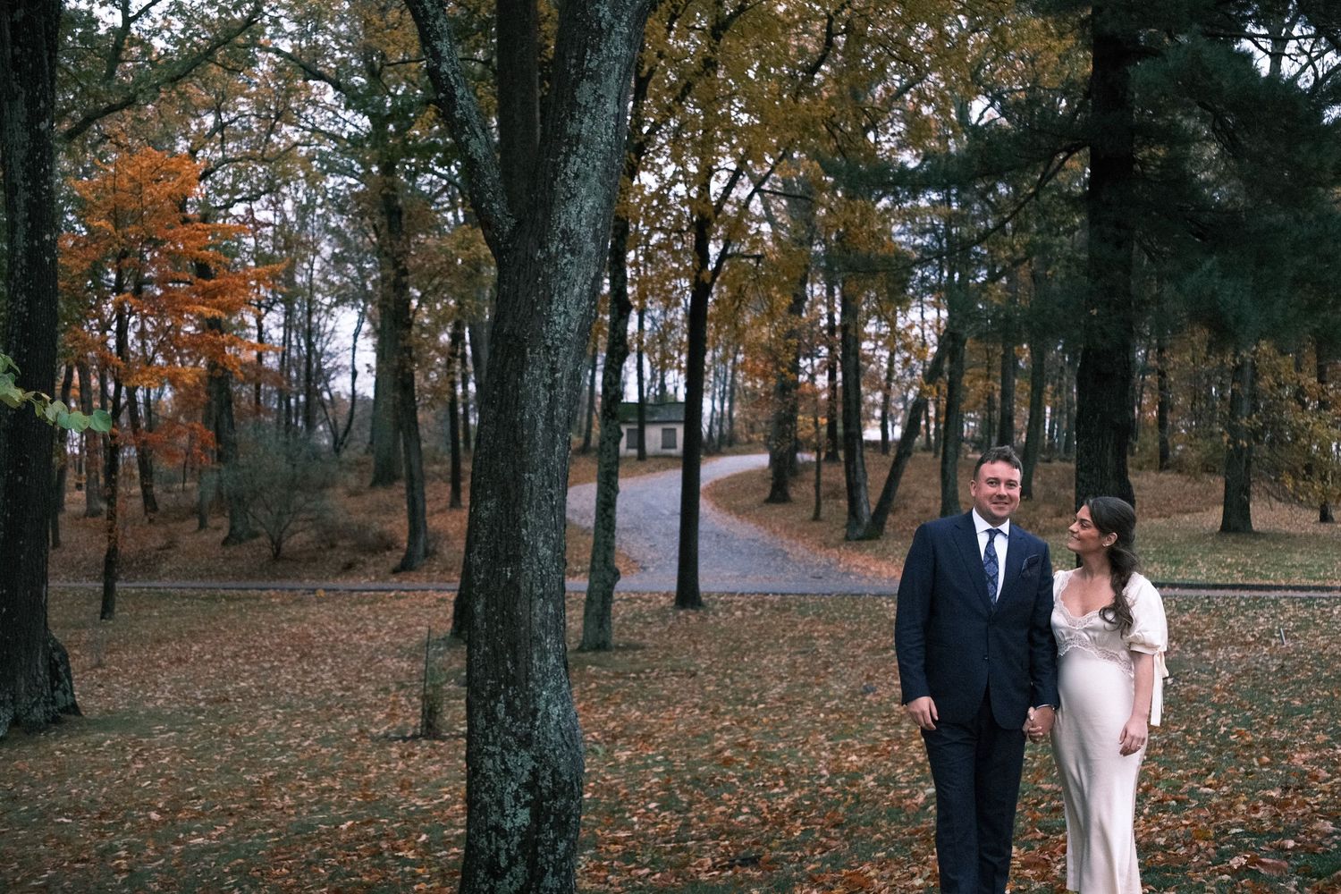 Couple in wedding attire walking through autumn forest with colorful foliage and a path leading to a house.