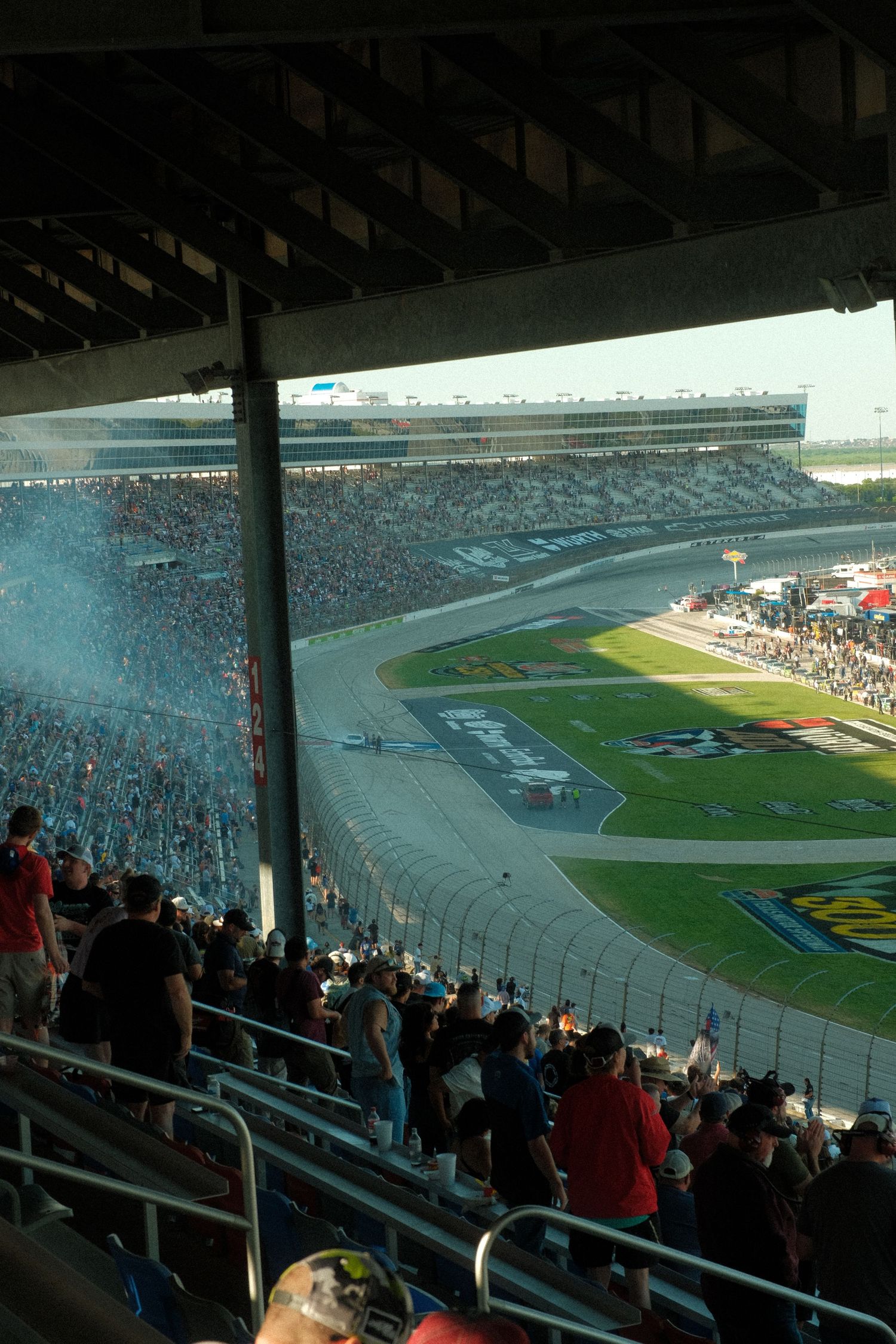 Packed NASCAR race stadium with fans watching cars on the oval track, smoke rising from the speedway under a hazy sky.
