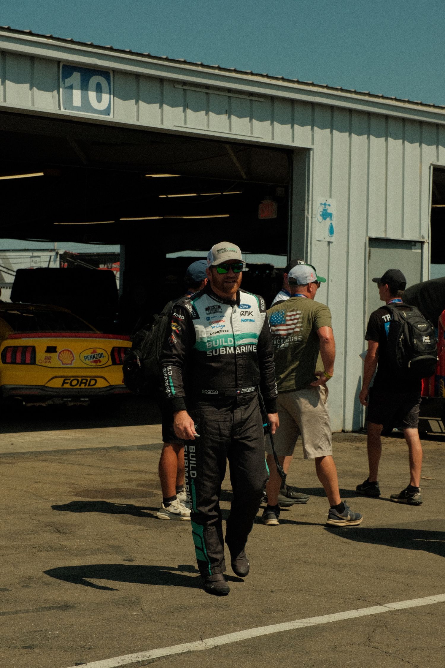 Racing driver in black firesuit walks past pit garage number 10 at a motorsport event with crew members and Ford race car.