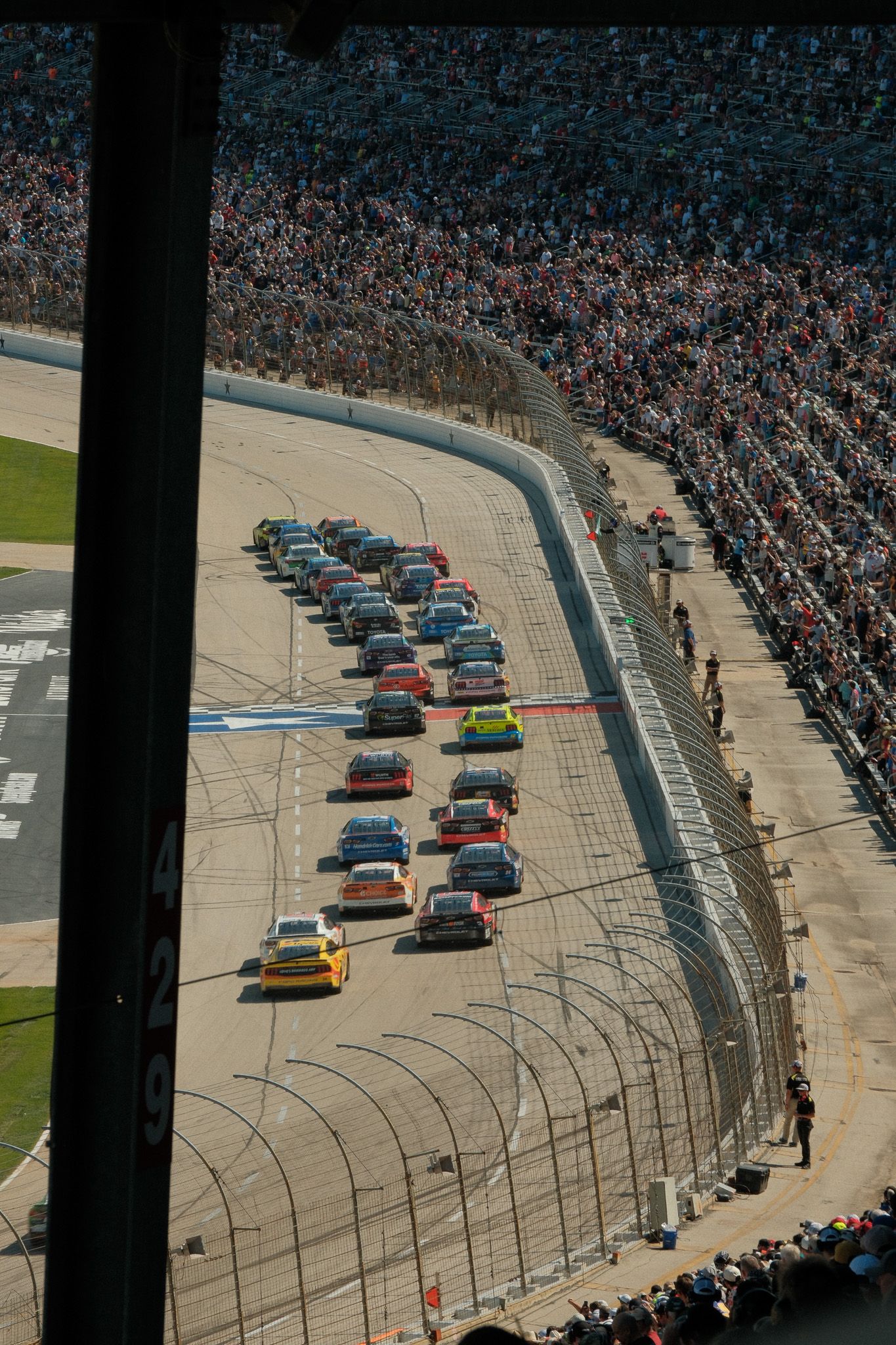 Aerial view of NASCAR race cars lined up on a banked oval track with packed grandstands full of spectators.
