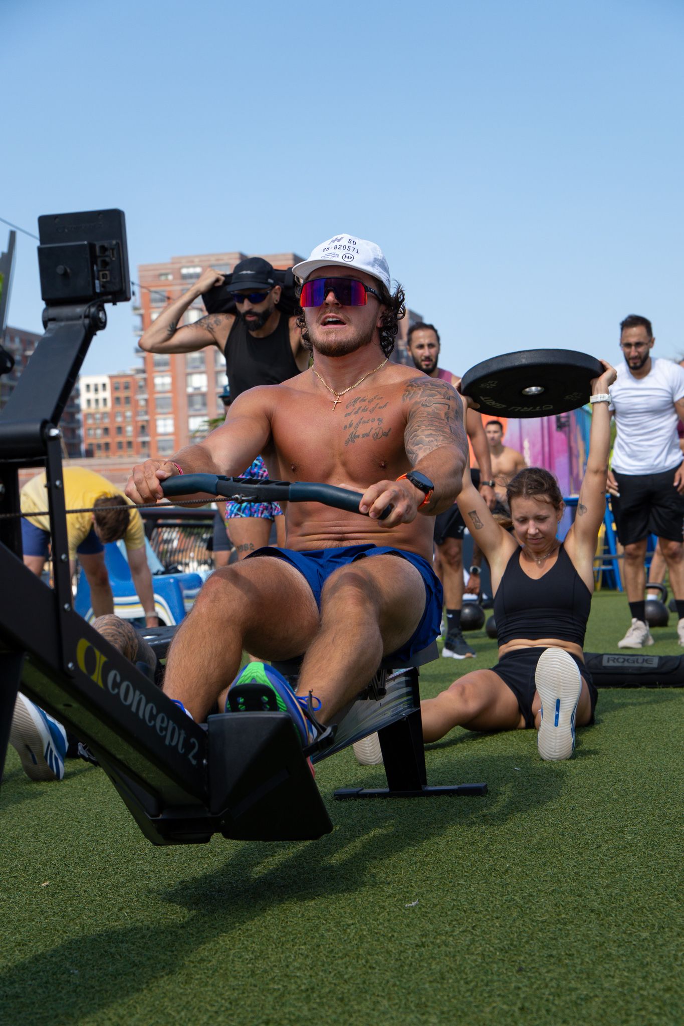 Muscular shirtless man using rowing machine outdoors during group fitness workout on turf area.
