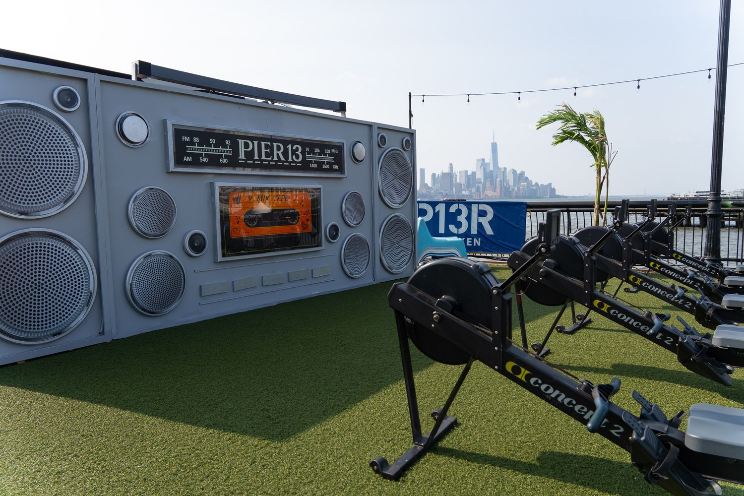 Outdoor fitness area at Pier 13 featuring rowing machines on artificial turf with a giant boombox backdrop and NYC skyline.