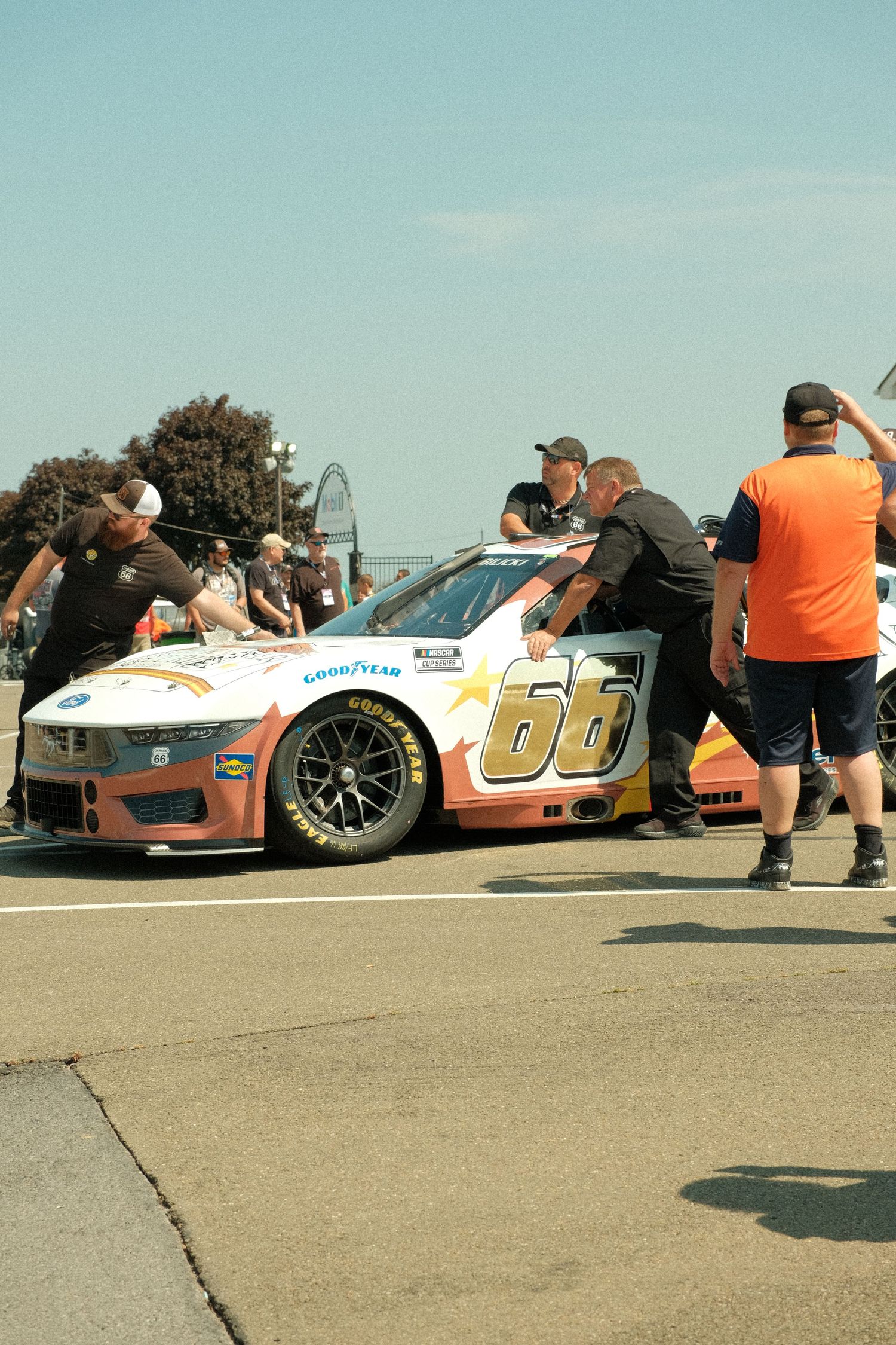 Crew members pushing white NASCAR #66 race car through pit road at sunny outdoor speedway event.