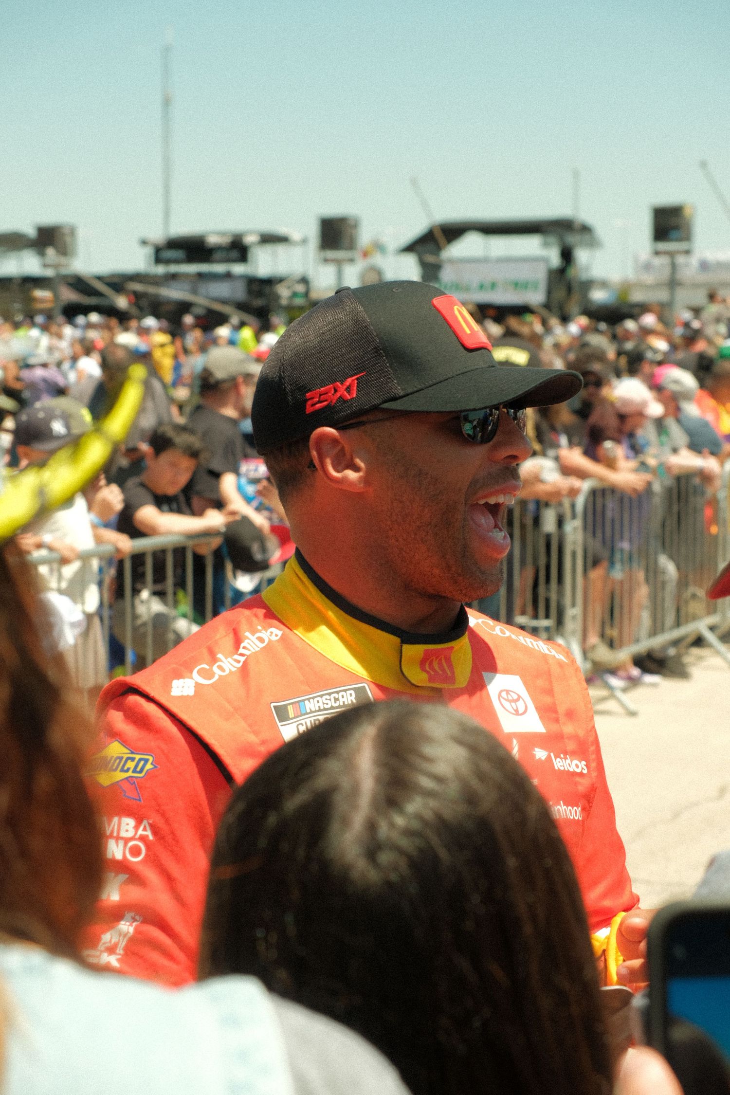 NASCAR driver Bubba Wallace in red Coca-Cola racing suit and black cap interacting with fans at a crowded race event.