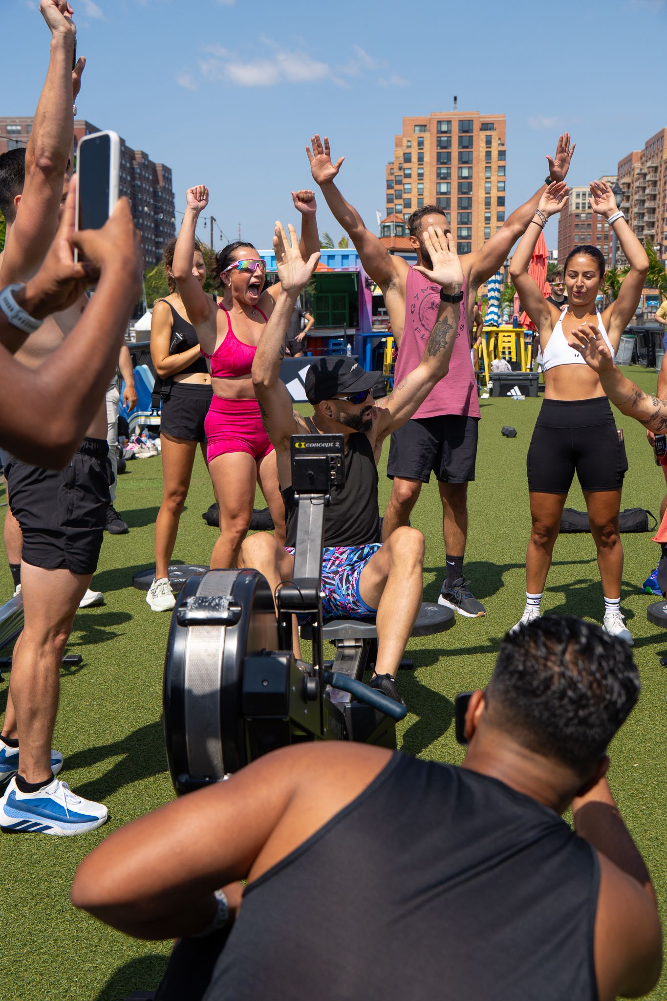 Group fitness class outdoors with participants cheering around a rowing machine on green turf in an urban setting.