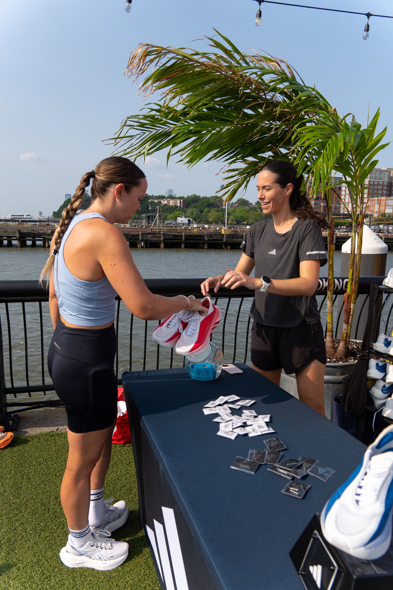 Two women at an Adidas outdoor event table by the waterfront, exchanging running shoes on a sunny day.