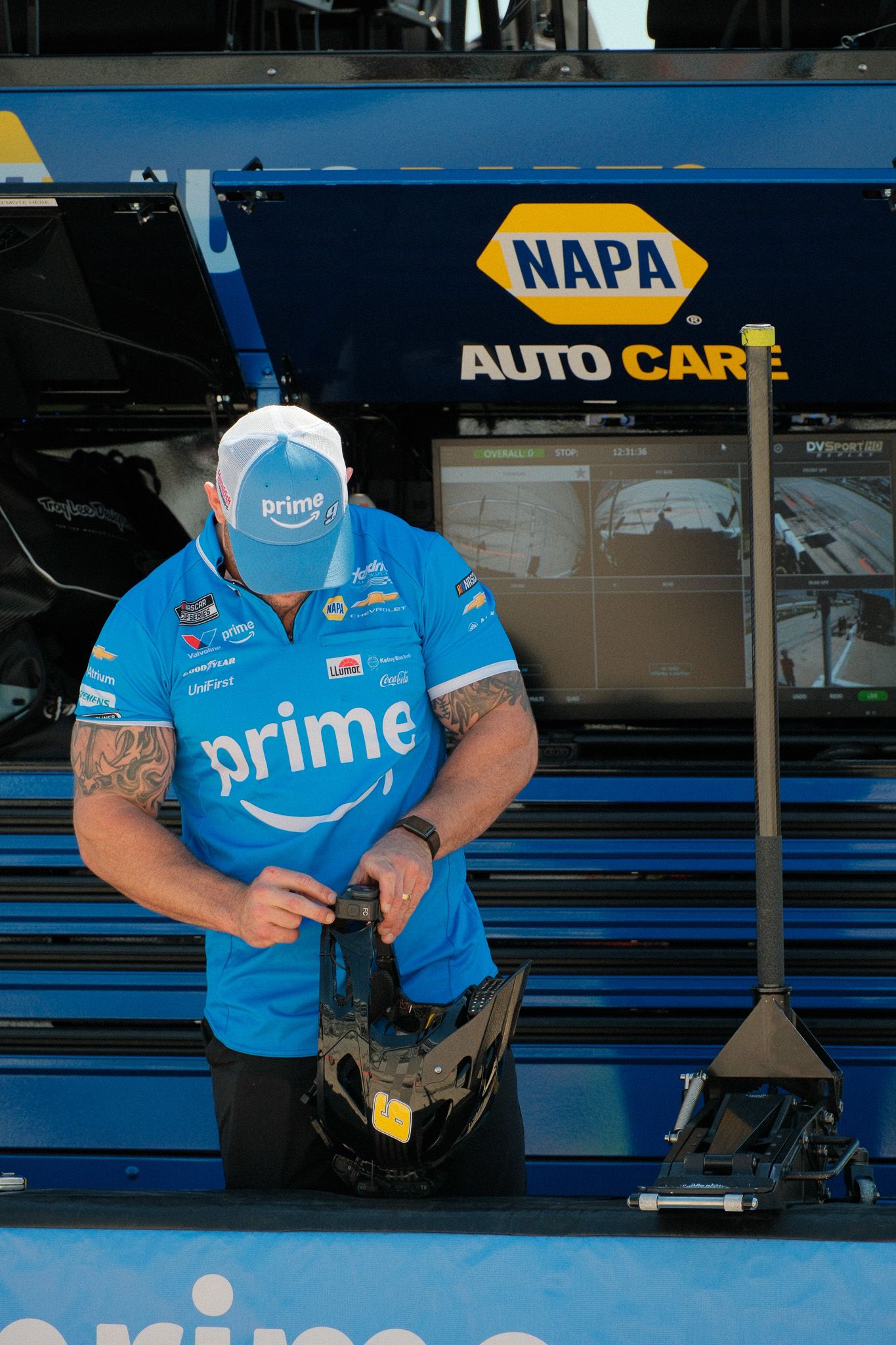 NASCAR Prime-sponsored crew member in blue uniform handles equipment near NAPA Auto Care hauler in pit area.