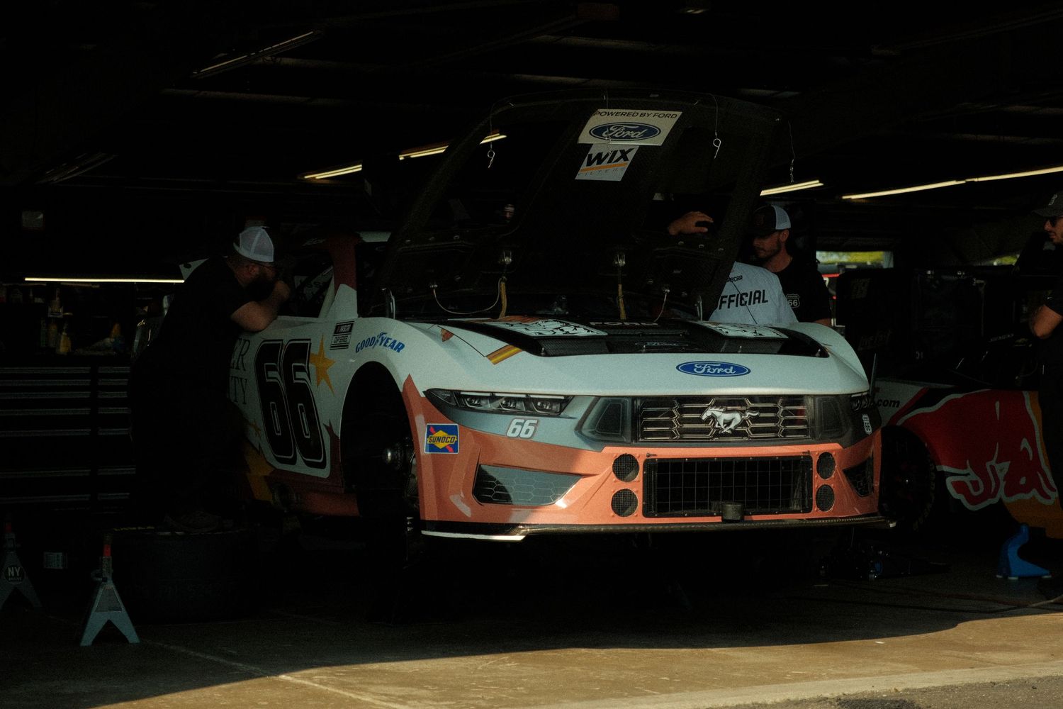 NASCAR Ford Mustang number 66 in a dark pit garage with crew members working nearby.