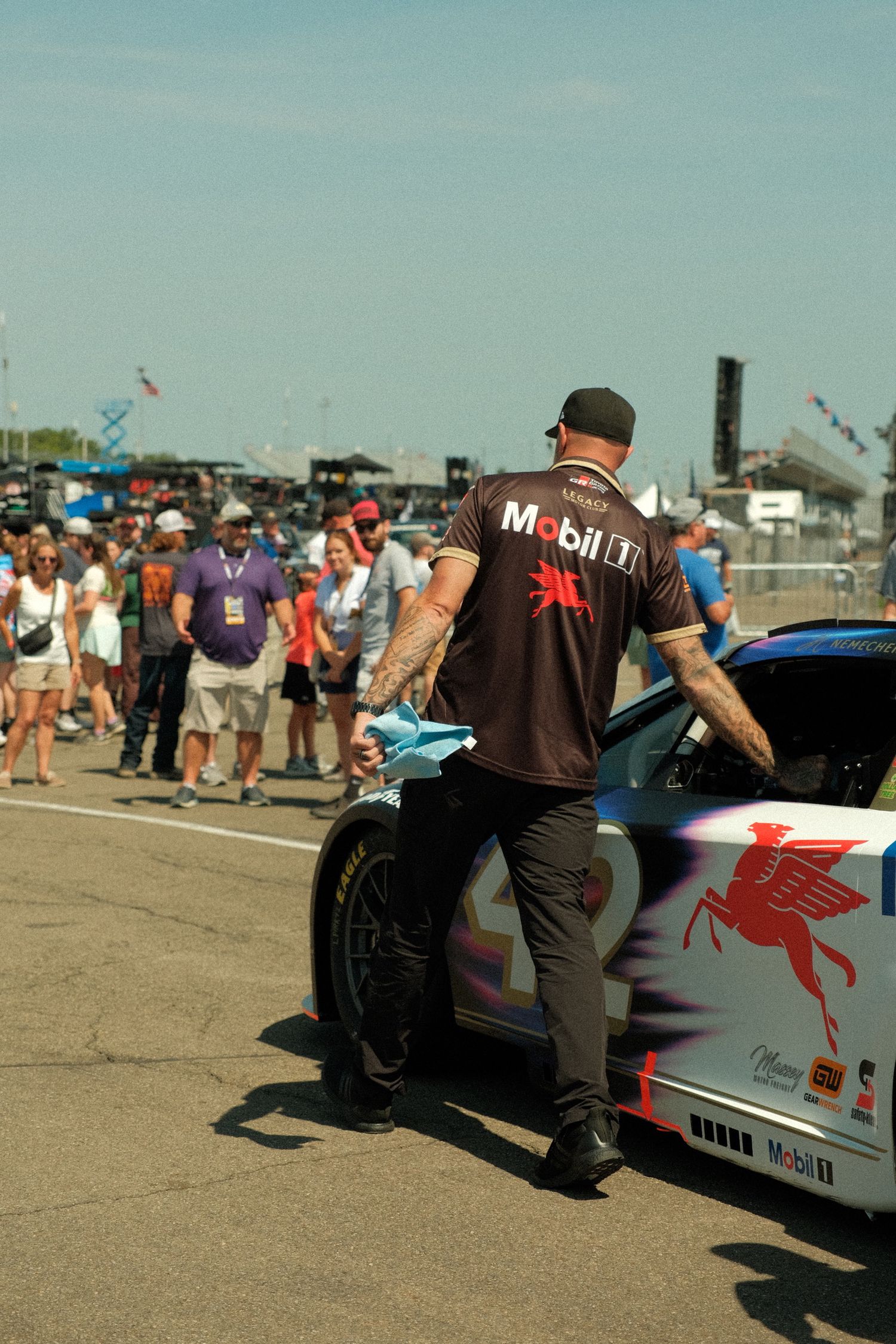 NASCAR crew member in Mobil 1 shirt tends to Pegasus-themed race car on pit road at crowded speedway.