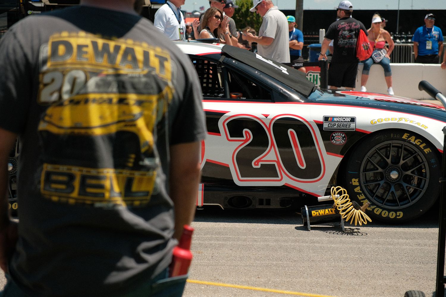 A fan wearing a DeWalt Christopher Bell #20 NASCAR shirt stands near the race car in the pit area.