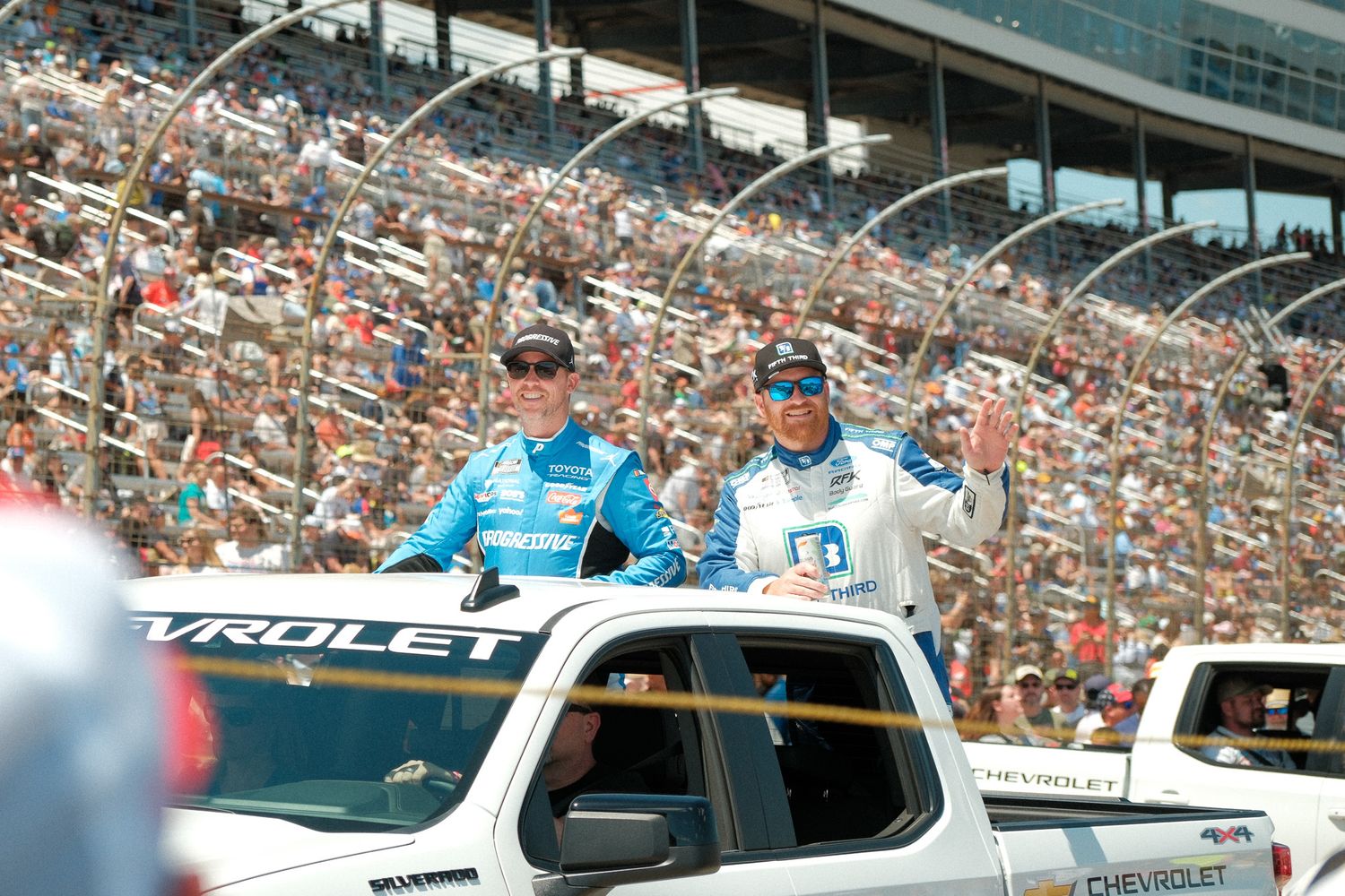 Two NASCAR drivers wave to fans from a Chevrolet truck during a pre-race driver parade.