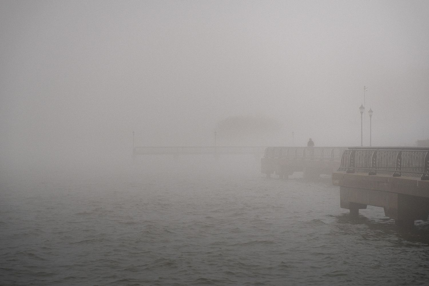Dense morning fog obscuring concrete pier and waterfront structures over calm gray harbor waters Hoboken, NJ.