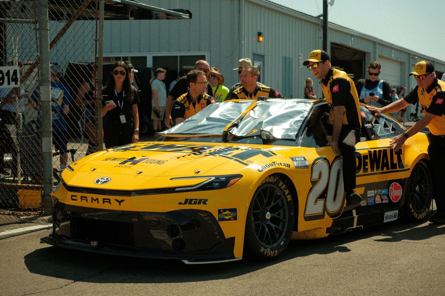 NASCAR #20 DeWalt Toyota Camry race car being pushed through pit road by crew members at a sunny race track.