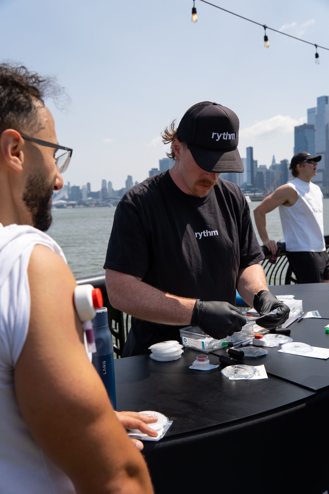 Medical professional in Rythmn cap administering vaccine outdoors with NYC skyline backdrop on sunny day.