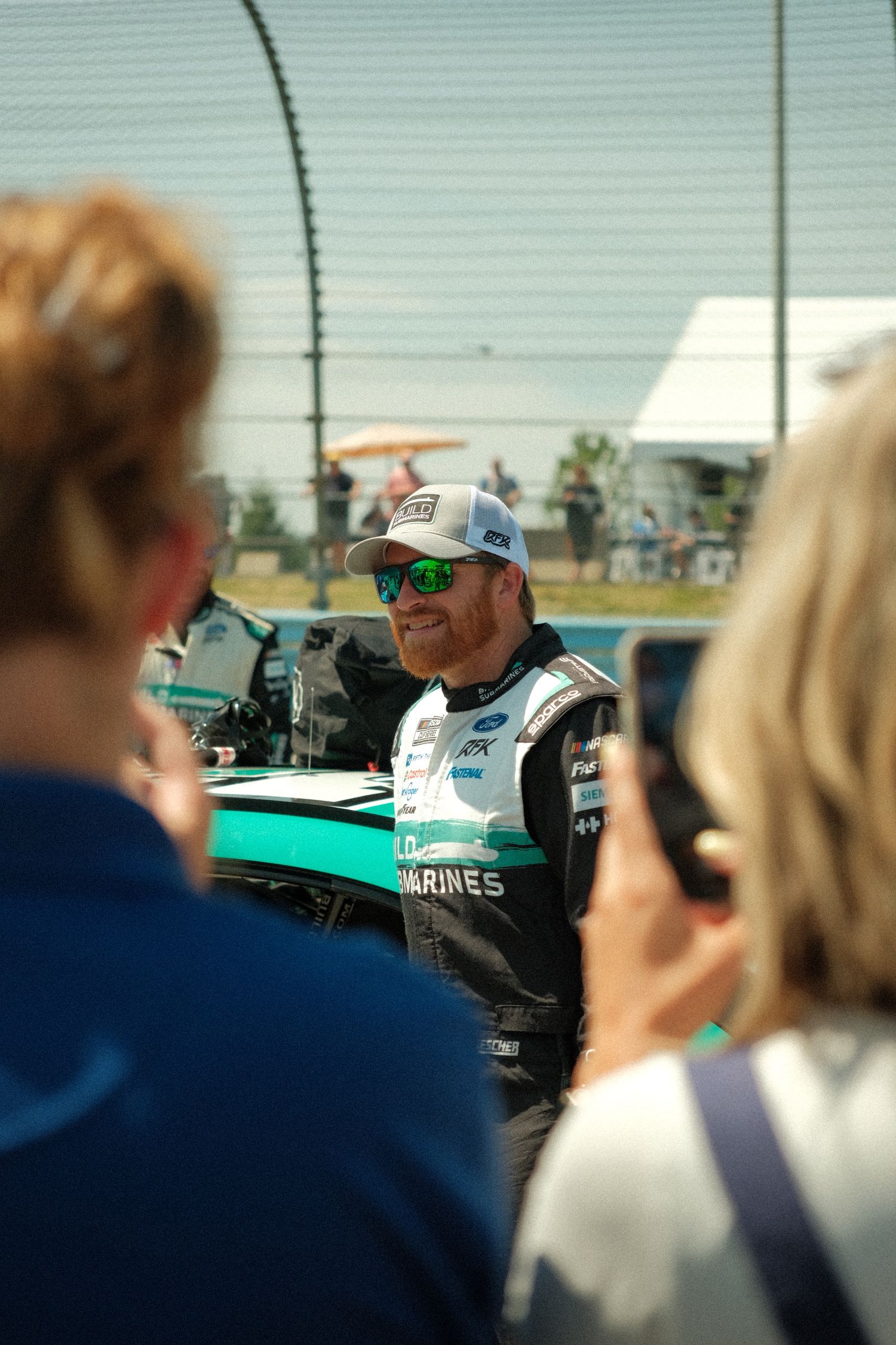 NASCAR driver in racing suit with green sunglasses and cap interacts with fans at a sunny race event.