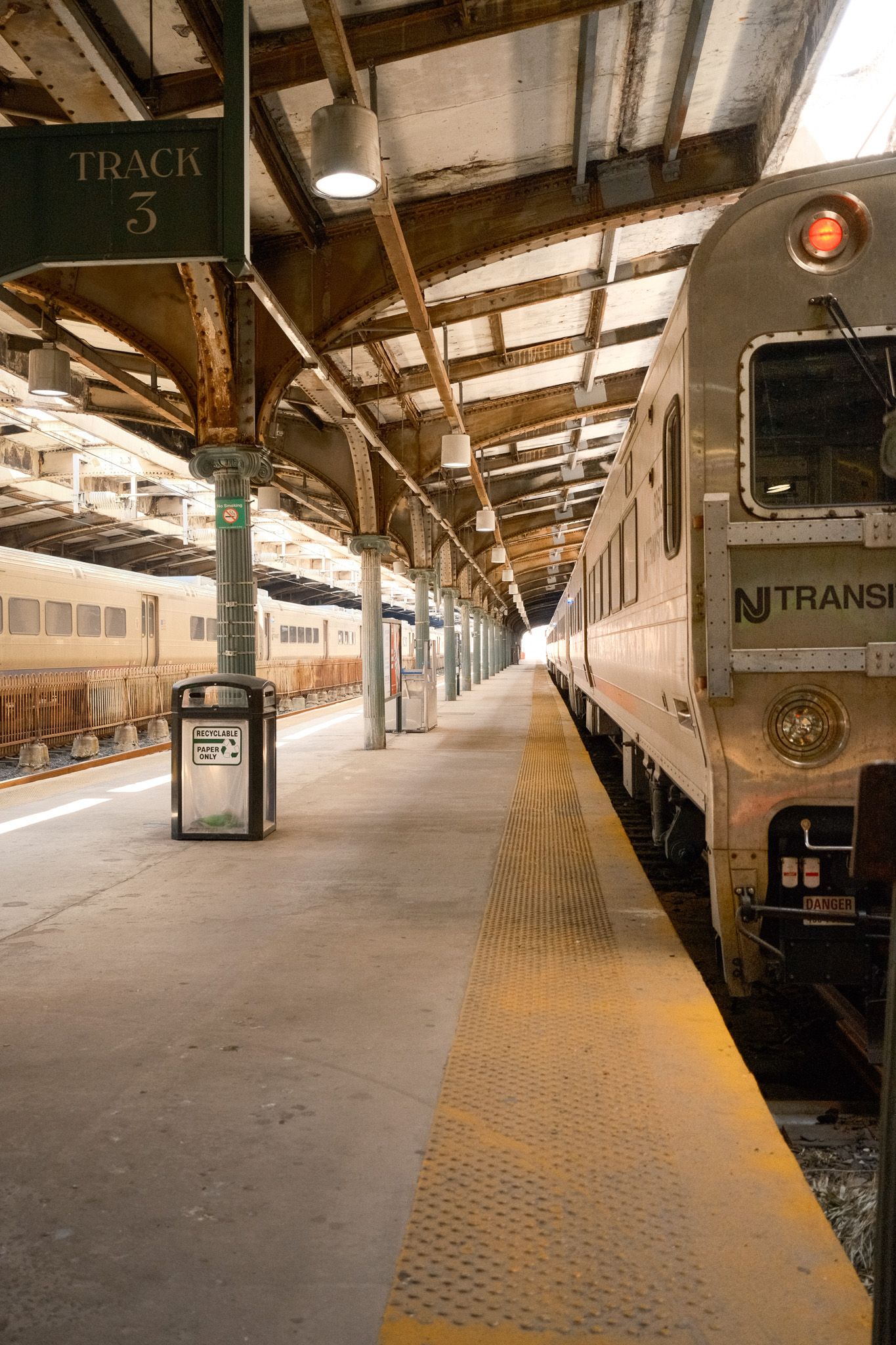 NJ Transit train parked at Track 3 platform inside Hoboken Terminal station with industrial ceiling.