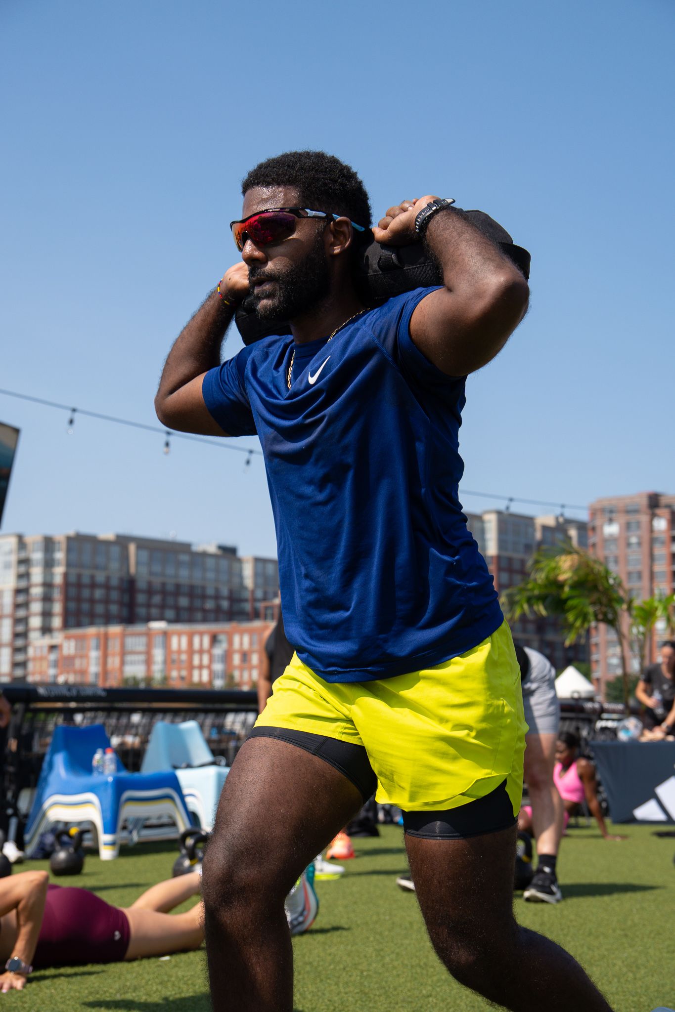Athletic man in blue Nike shirt and yellow shorts exercises outdoors at urban fitness event on sunny day.