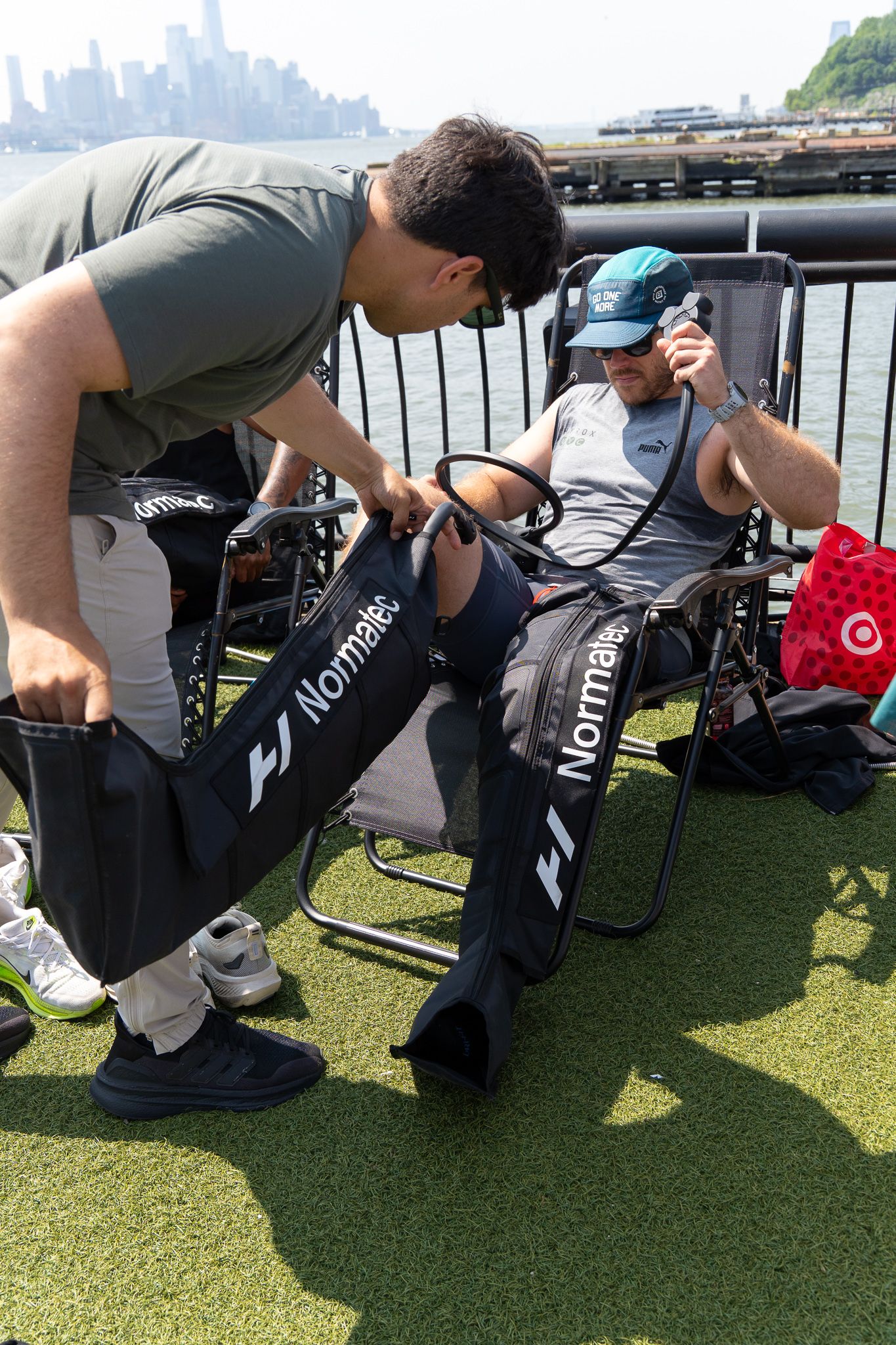 Two men using Normatec recovery boots outdoors by a waterfront on a sunny day.