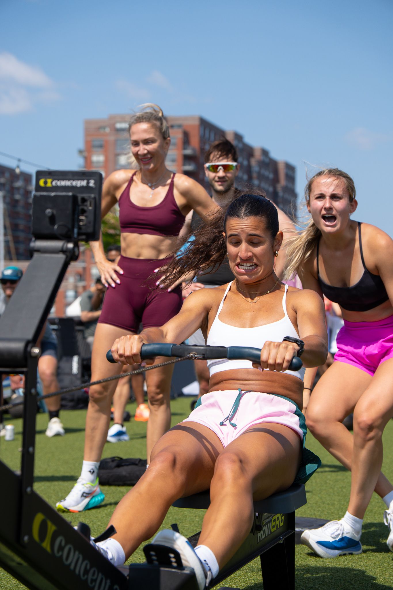 Woman intensely rowing on Concept2 machine outdoors while teammates cheer her on.