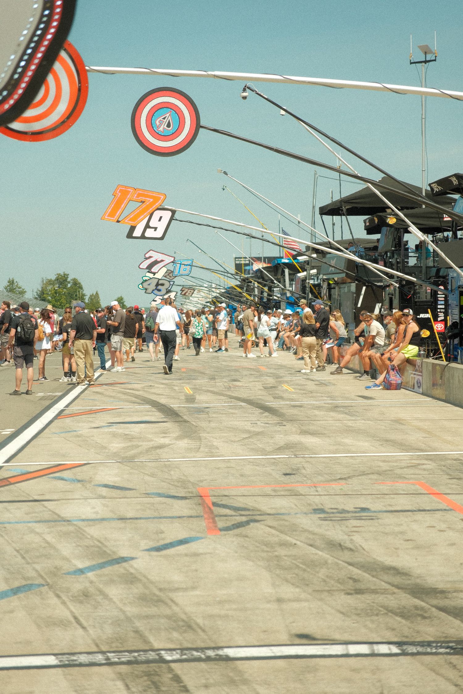 NASCAR pit road scene with team signs, crew members, and fans lining the track under a clear blue sky.