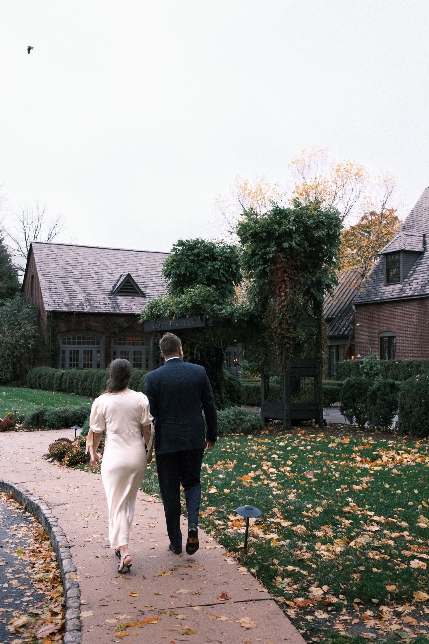 Couple walking hand in hand along a brick path toward an ivy-covered stone manor surrounded by autumn foliage.