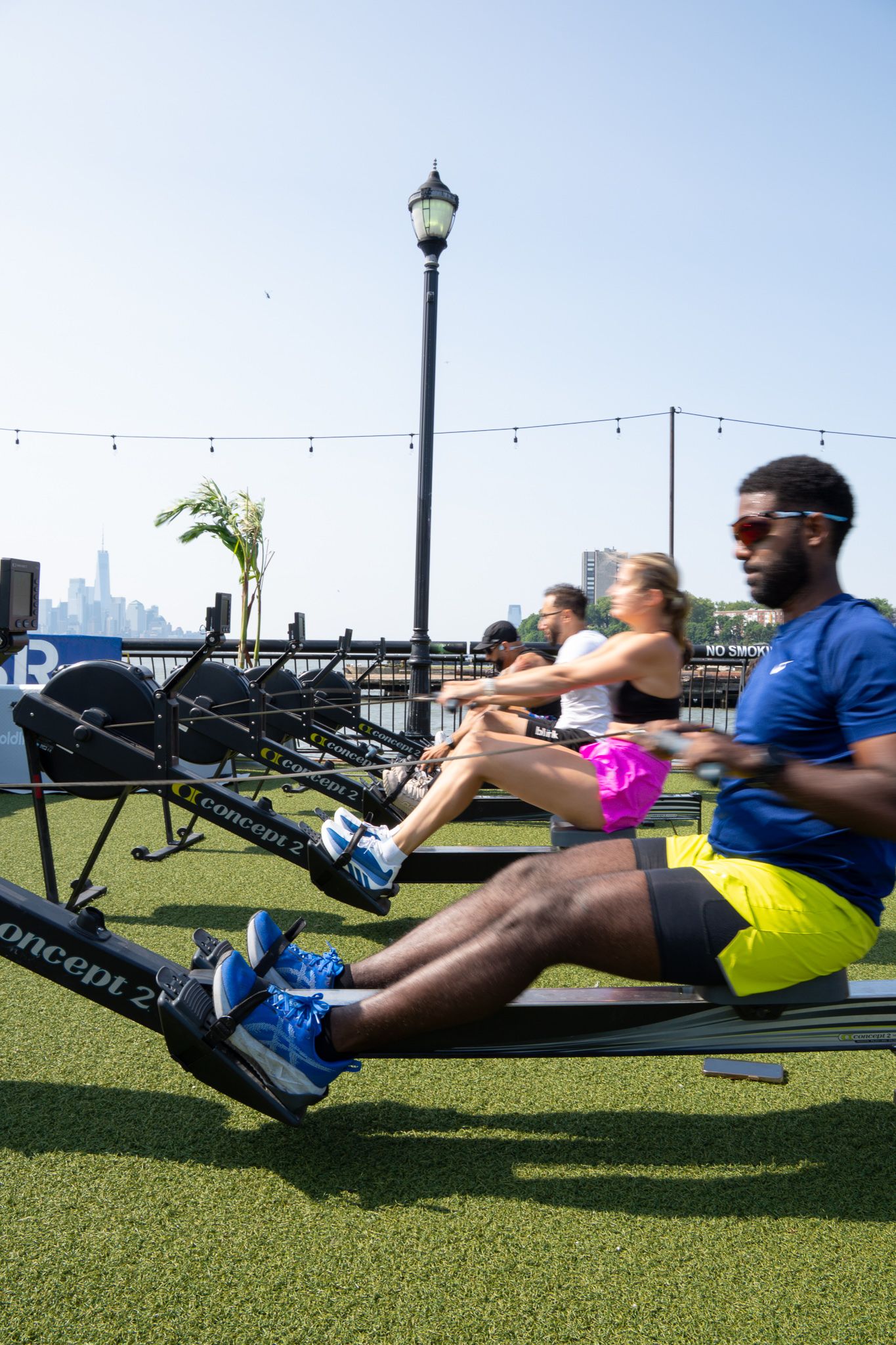 Group fitness class doing rowing machine exercises outdoors on turf with city skyline in background on sunny day.