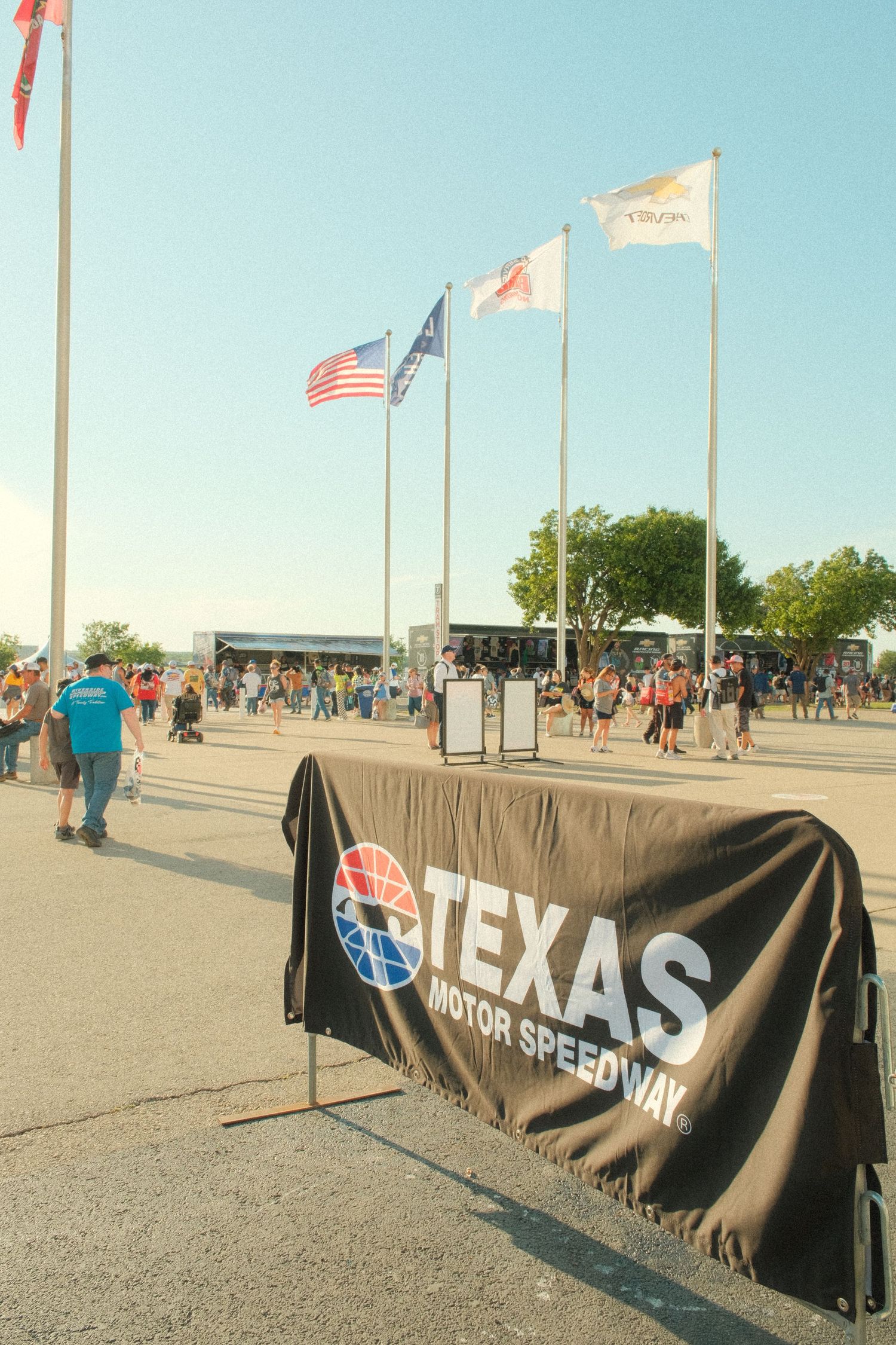 Texas Motor Speedway banner at entrance with American flags flying on poles, crowds of racing fans gathering on sunny day.